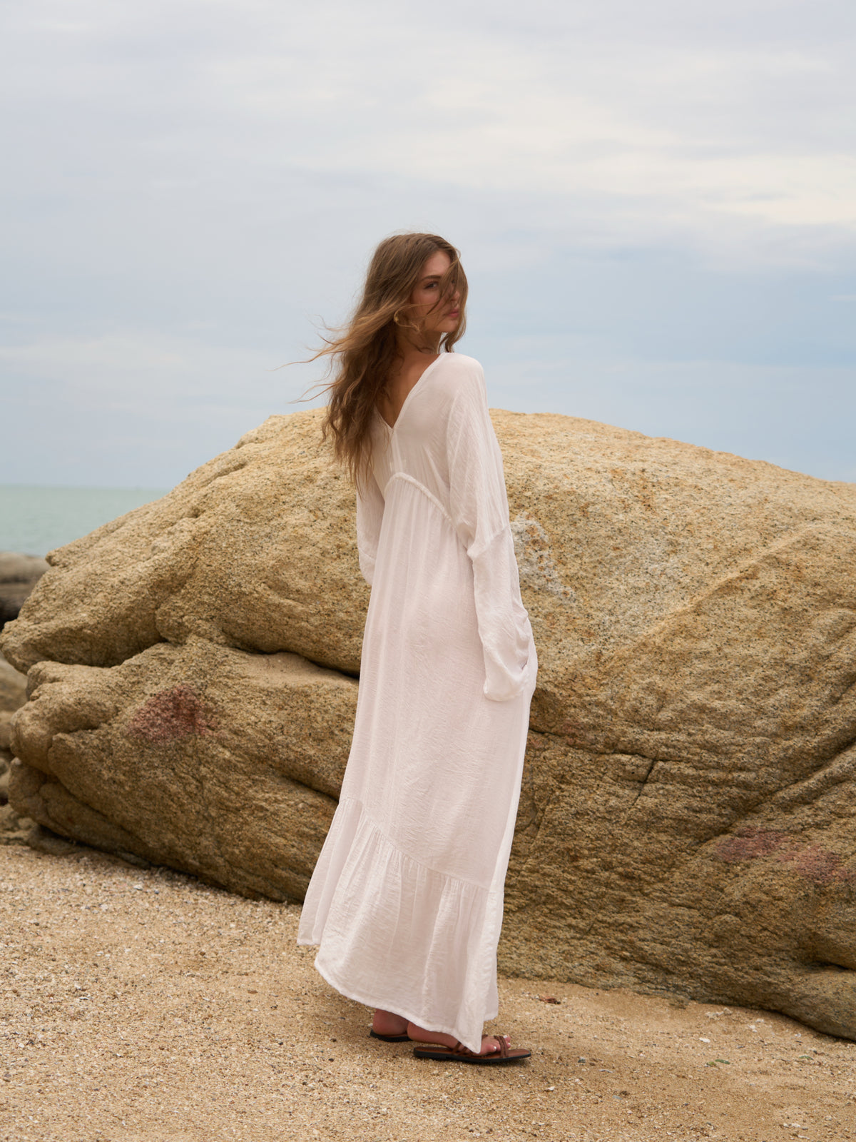 A woman wearing a long white maxi dress stands on a sandy beach next to large tan boulders under an overcast sky.