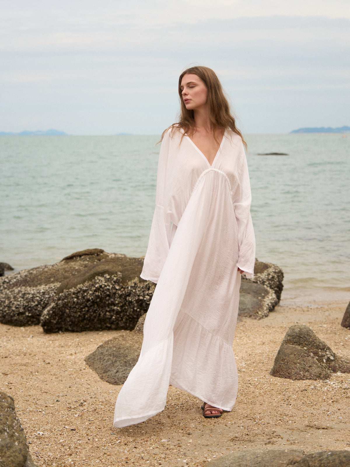 A woman wearing a flowing white long-sleeve maxi cover-up stands on a sandy beach with rocks and the ocean in the background.