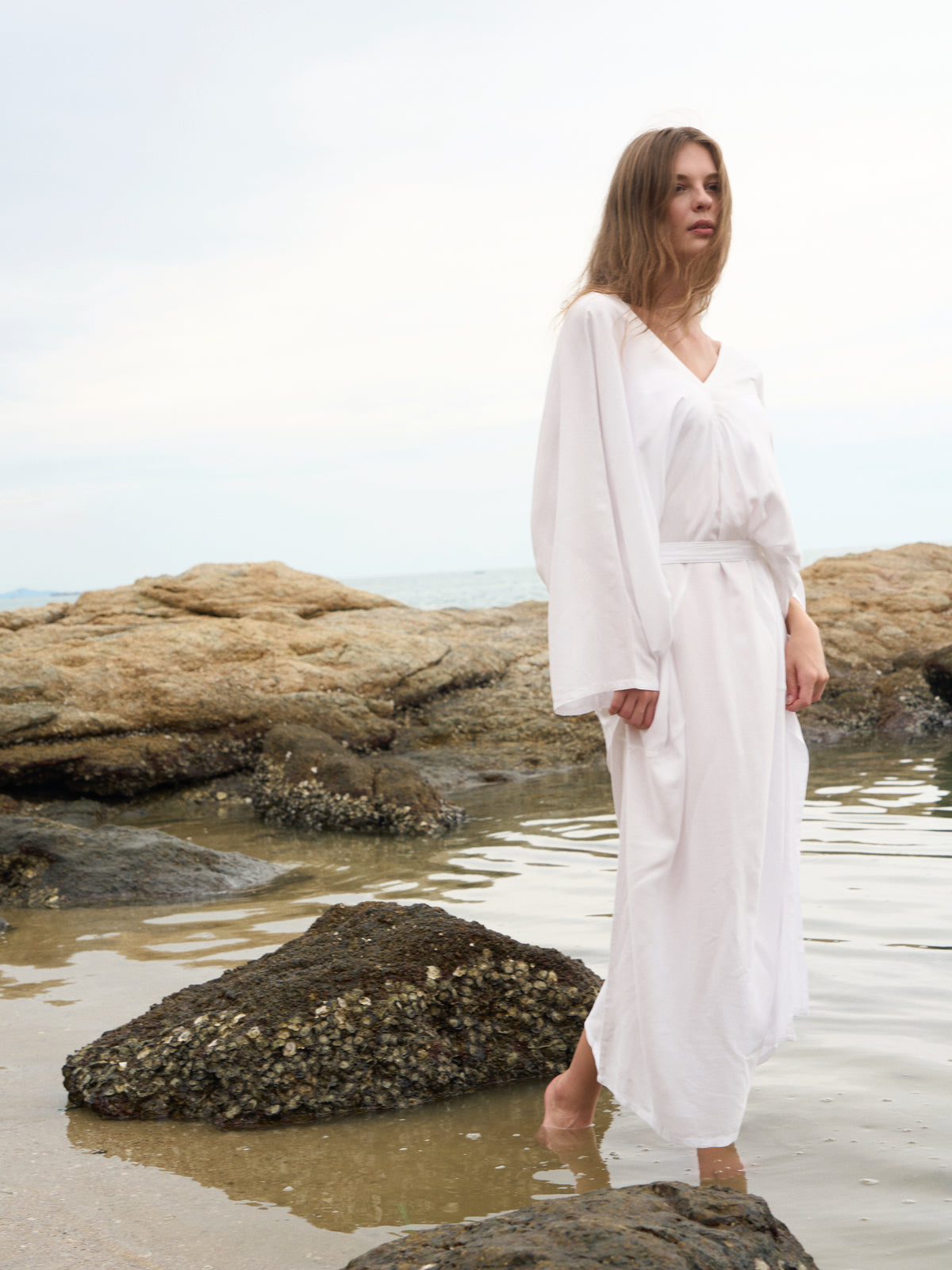 A woman wearing a white long-sleeve maxi robe stands barefoot on wet rocks at the beach, with ocean water and tan rocks in the background.