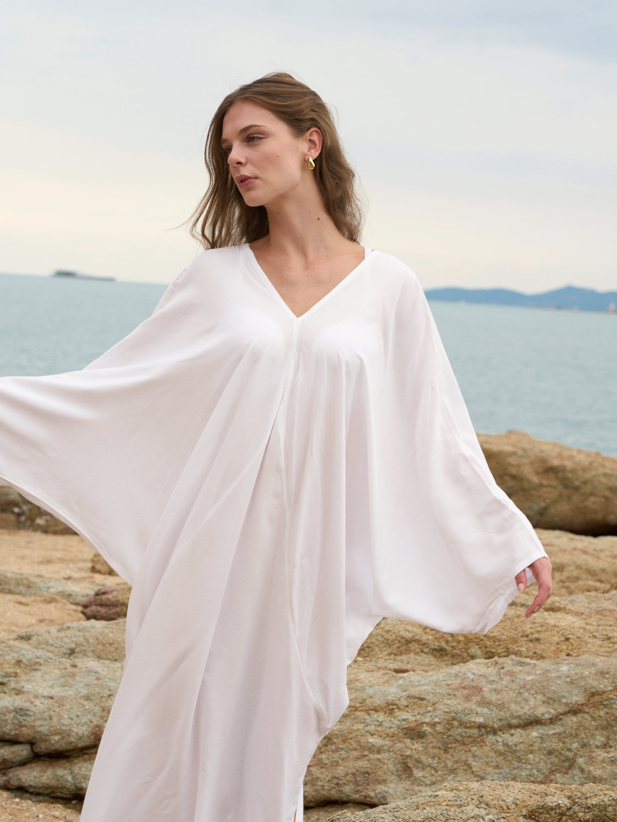 A woman wearing a flowing white caftan-style cover-up with wide draped sleeves, standing on rocky shoreline with the sea and sky in the background.