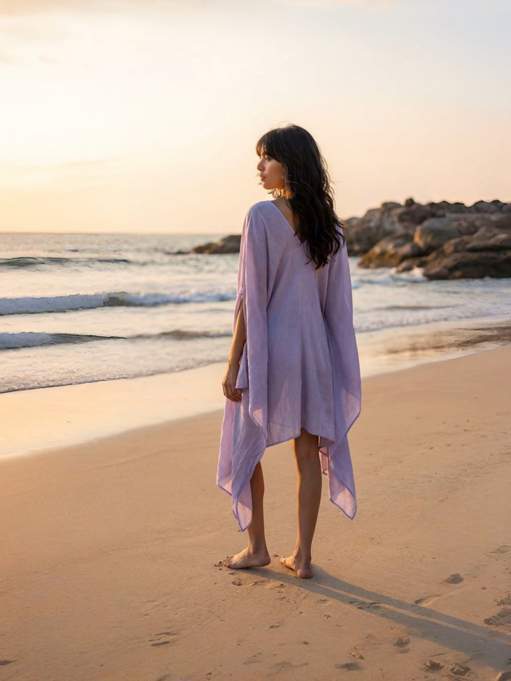 A woman stands on a sandy beach wearing a flowing lavender beach cover-up mini kaftan dress, with ocean waves and rocky shoreline in the background.