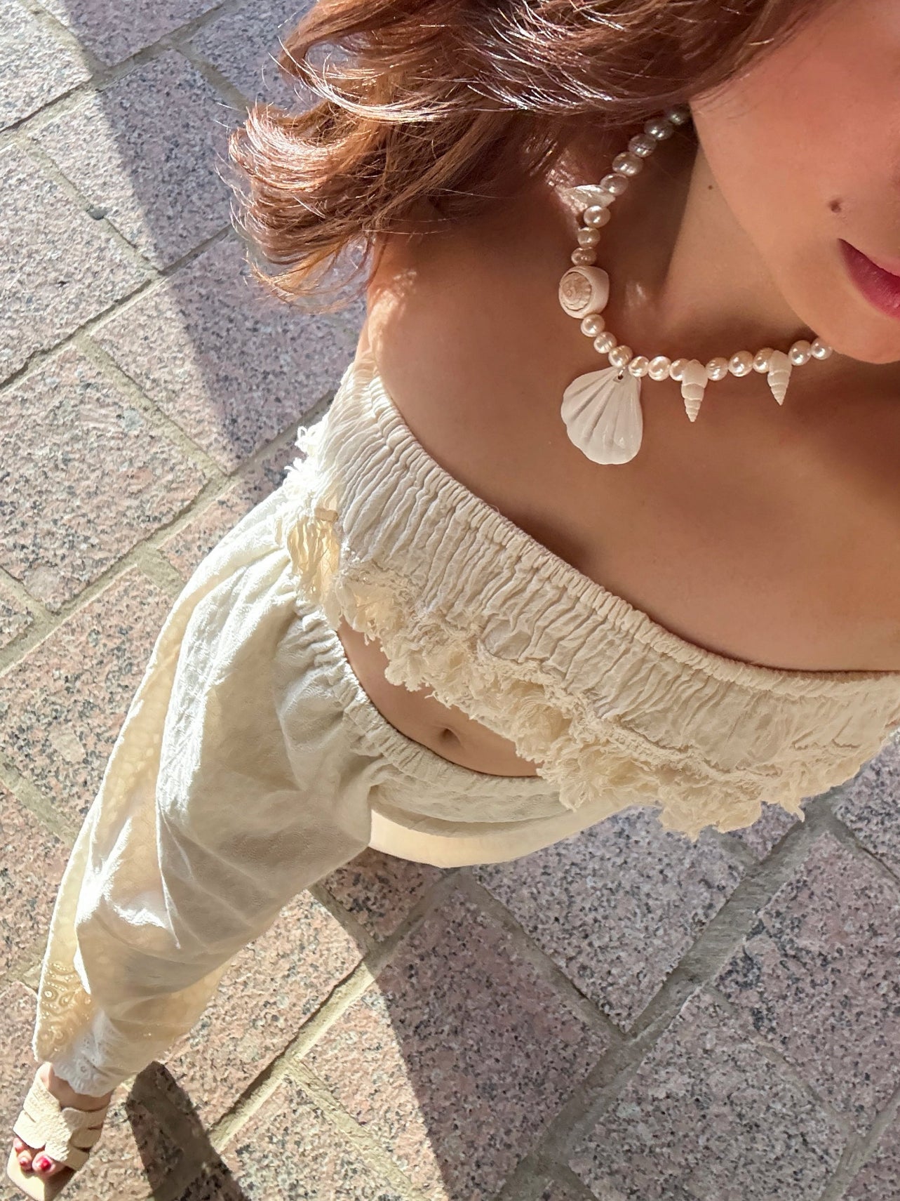 Person wearing a pearl seashell necklace and light-colored outfit on a stone pavement, boho style, beach accessories.