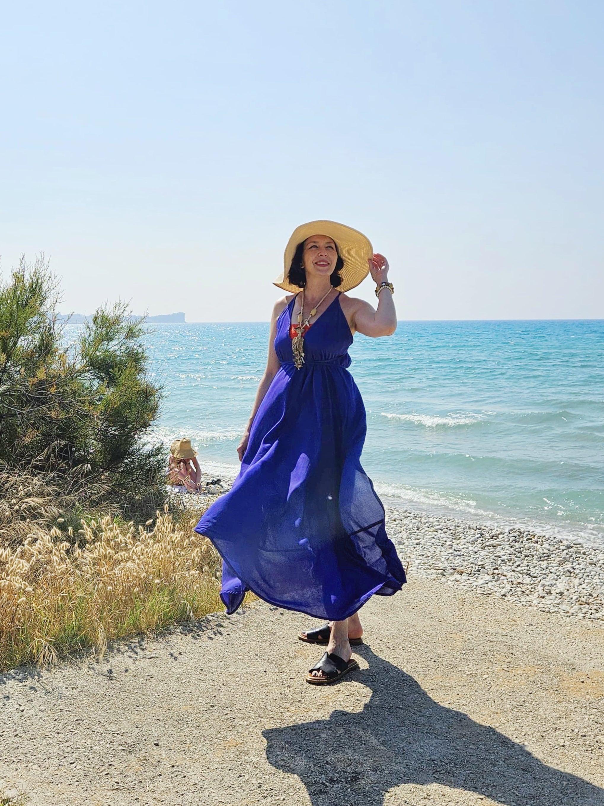 Woman wearing a flowy, hand-dyed purple blue halter maxi dress with open back and high slit, styled as beachy resort wear standing on a beach with ocean in the background such a beautiful vivid blue beach dress.