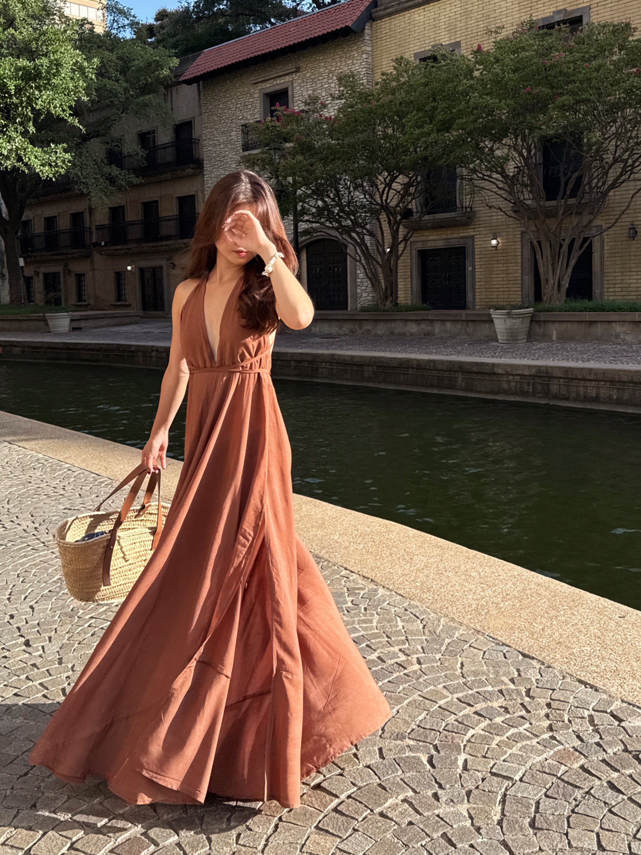 Woman in a long rust-colored maxi halter dress flowy dress standing by a canal with buildings and trees in the background.
