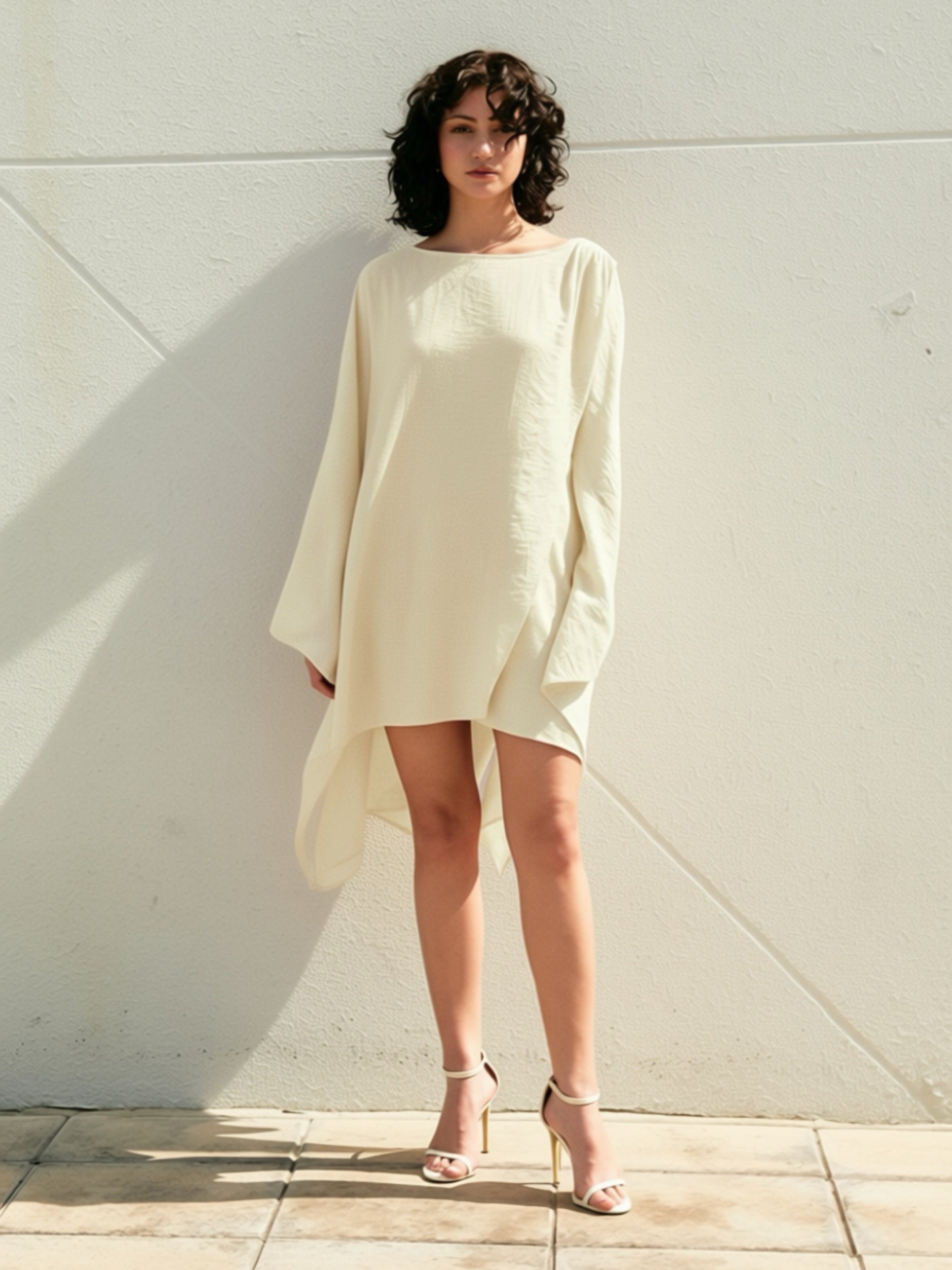 Woman wearing a white mini kaftan top beach cover up tunic, one size fits most, long sleeves with high-low hem, lightweight resort vacation wear, standing against a white wall in bobo style.