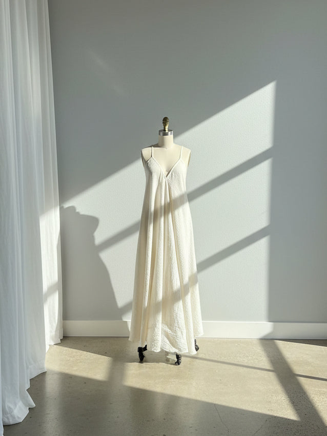 Ethically sourced organic cotton maxi dress displayed on a dress form, featuring a flowy A-line silhouette, deep V neckline, thin adjustable spaghetti straps, and a unique open back with tie detail. Handmade from raw, undyed cotton for a natural boho look, perfect as a beach dress or breezy summer weekend outfit.
