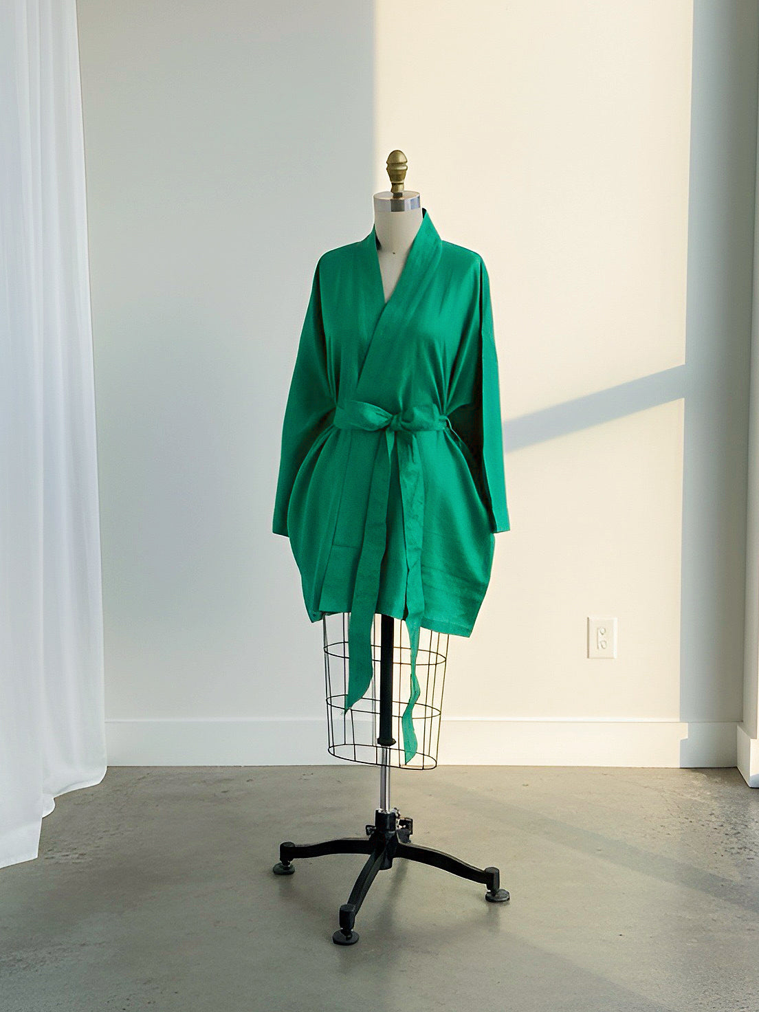 A bright green kimono-style robe displayed on a dress form mannequin with a black wire stand in a minimalist studio setting with a concrete floor and white walls.