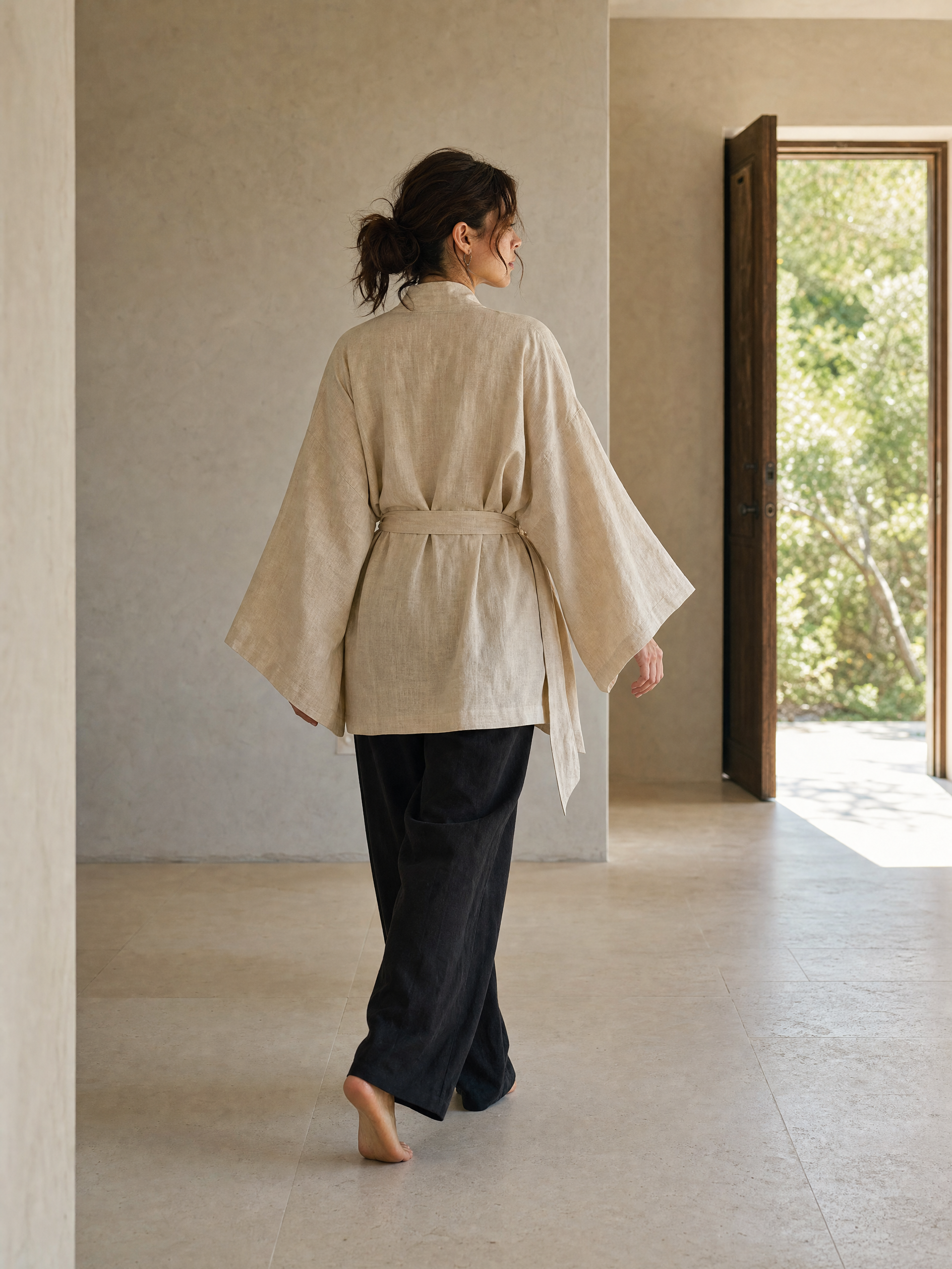 A woman is shown from behind wearing a light beige linen kimono-style top with wide sleeves and a tie belt, paired with dark wide-leg pants, walking barefoot in a minimalist interior with a doorway leading to greenery.