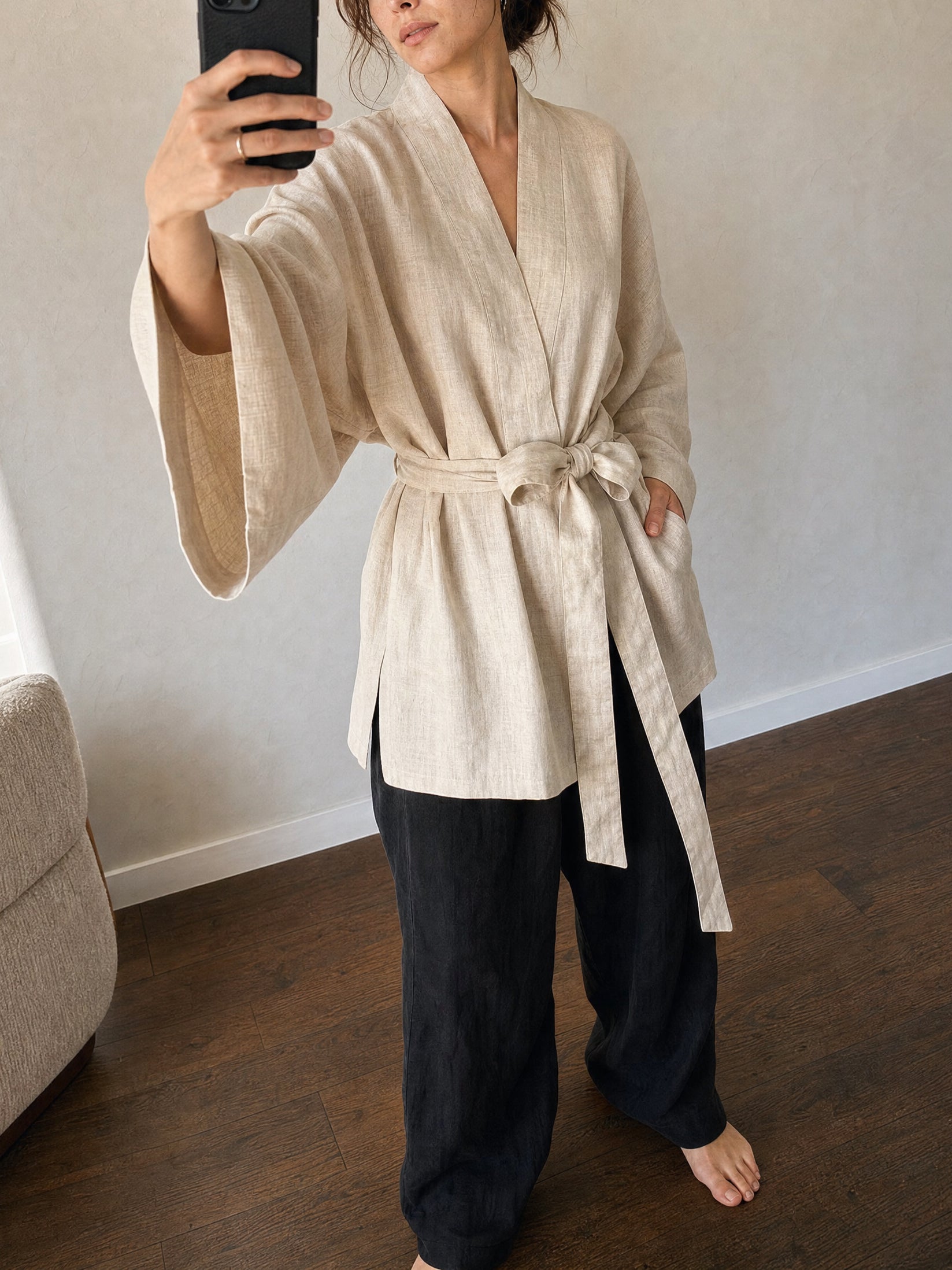 A woman takes a mirror selfie wearing a light beige cream  linen wrap robe with wide sleeves and a tie belt, paired with dark charcoal pants, in a minimalist room with wood floors.