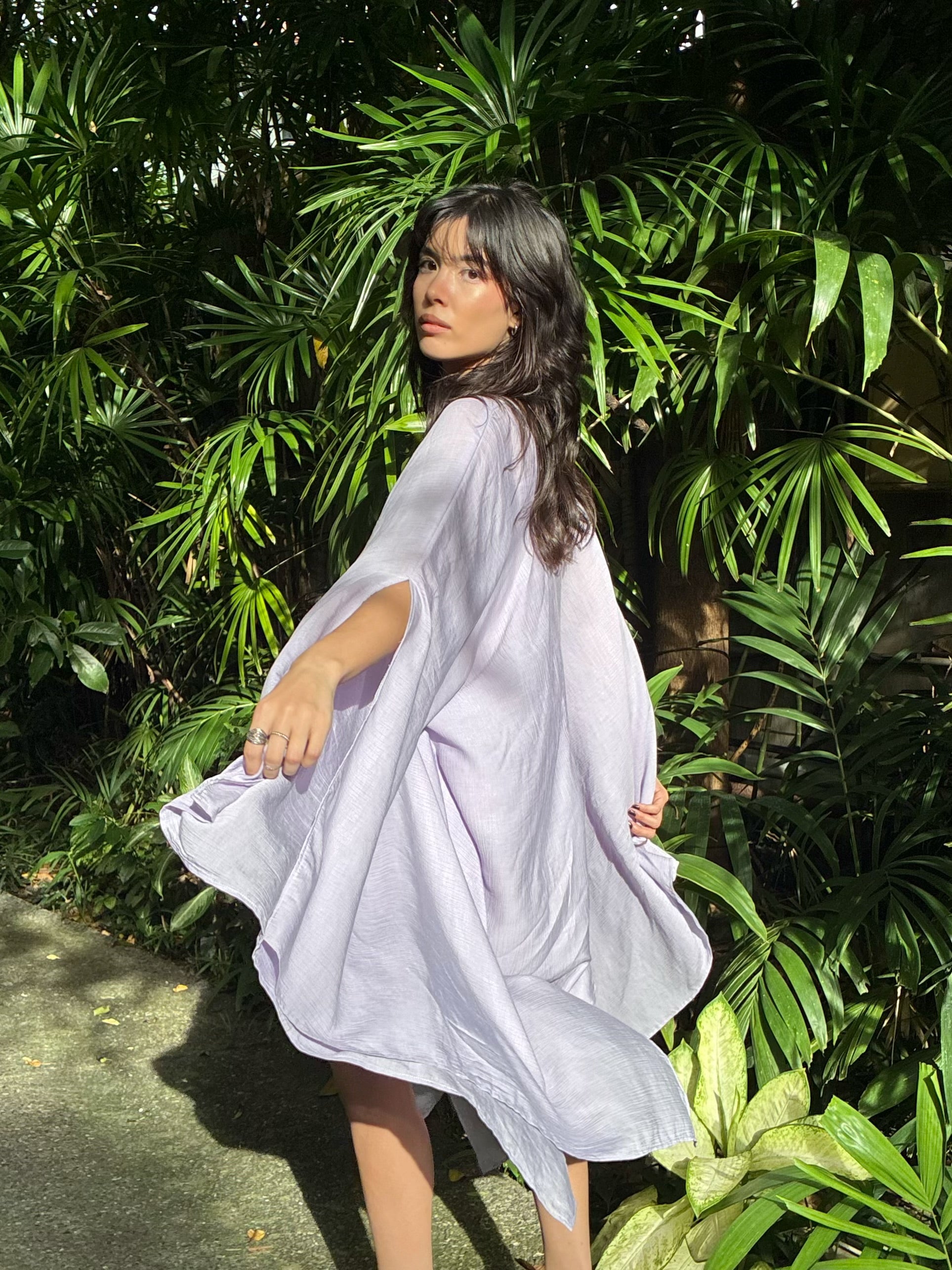 A woman wearing a flowing, loose-fitting light lavender or pale purple kaftan-style cover-up dress, standing outdoors in front of dense green tropical foliage. Perfect Kaftan Cover Up, flowy oversized beach cover up and resort top with deep V neckline for poolside lounging and cruise outfits