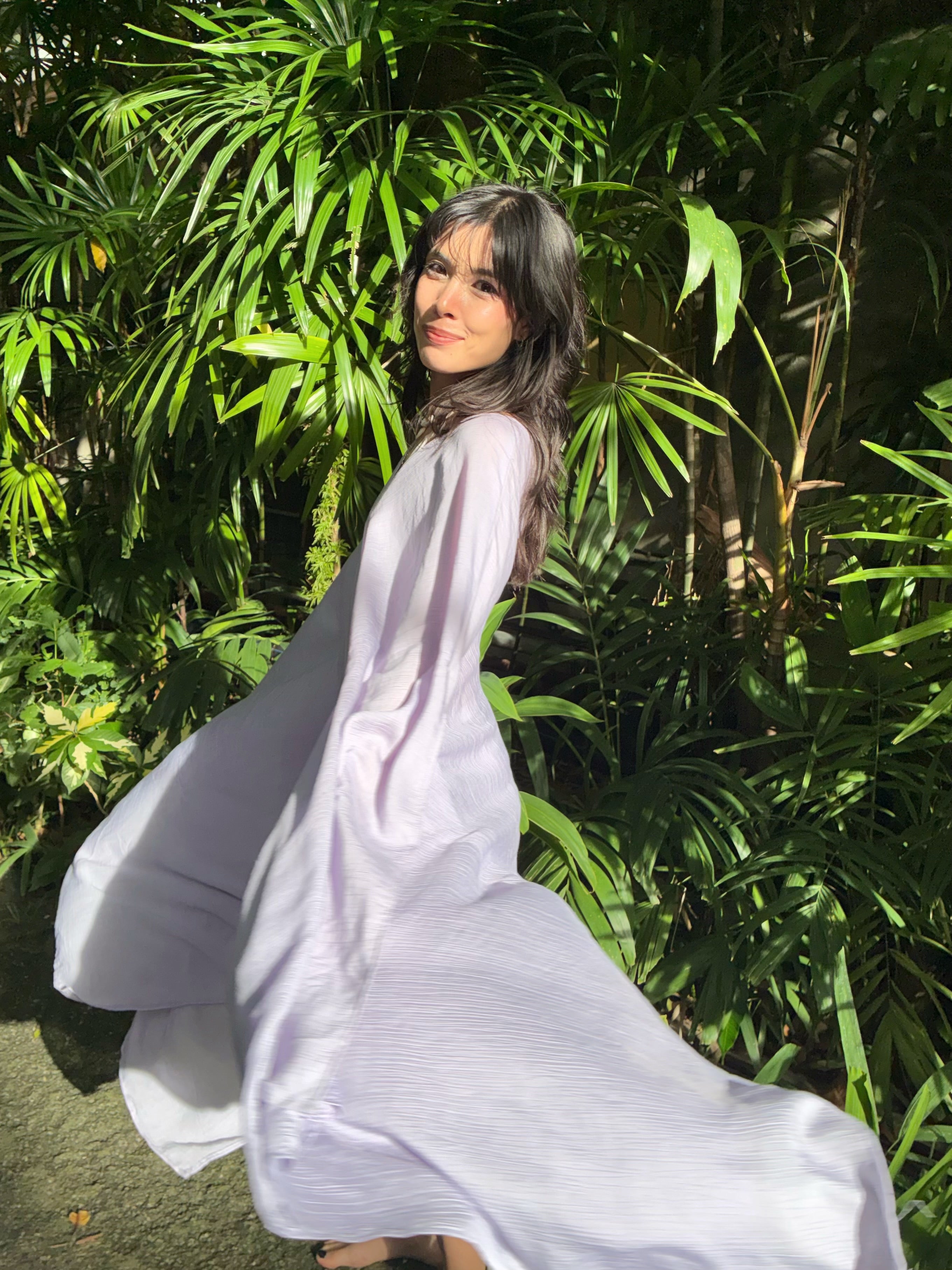 A woman wearing a flowing, light lavender mini kaftan dress stands outdoors in front of lush green tropical foliage, with the dress billowing in the wind.