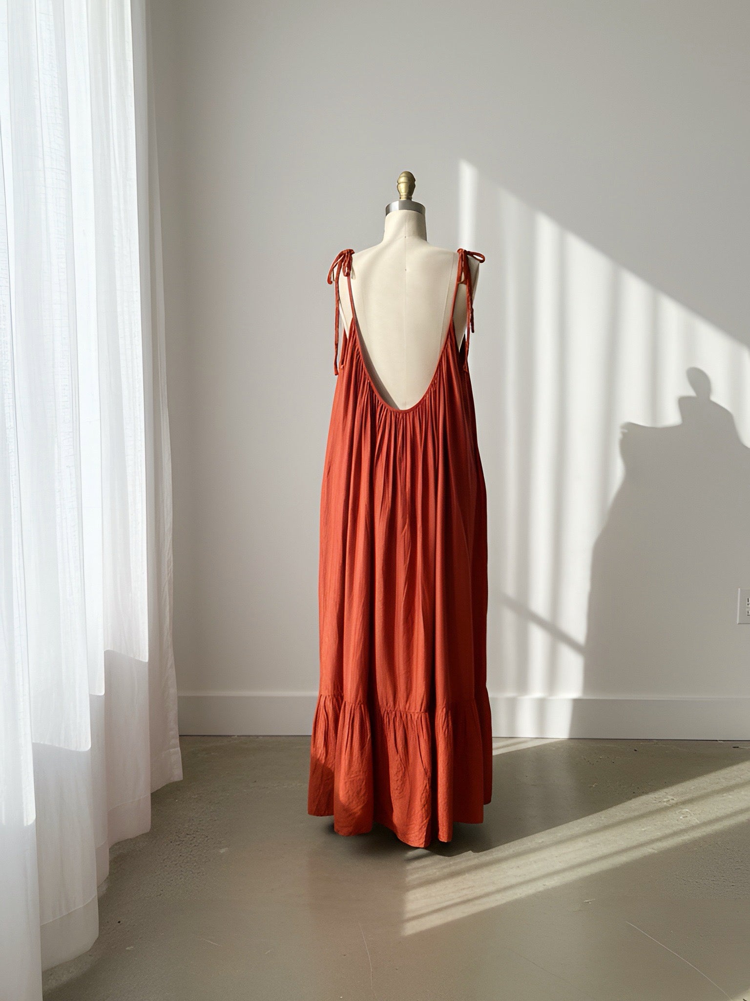 Burnt orange open back maxi dress on a mannequin against a white wall with sunlight casting shadows.