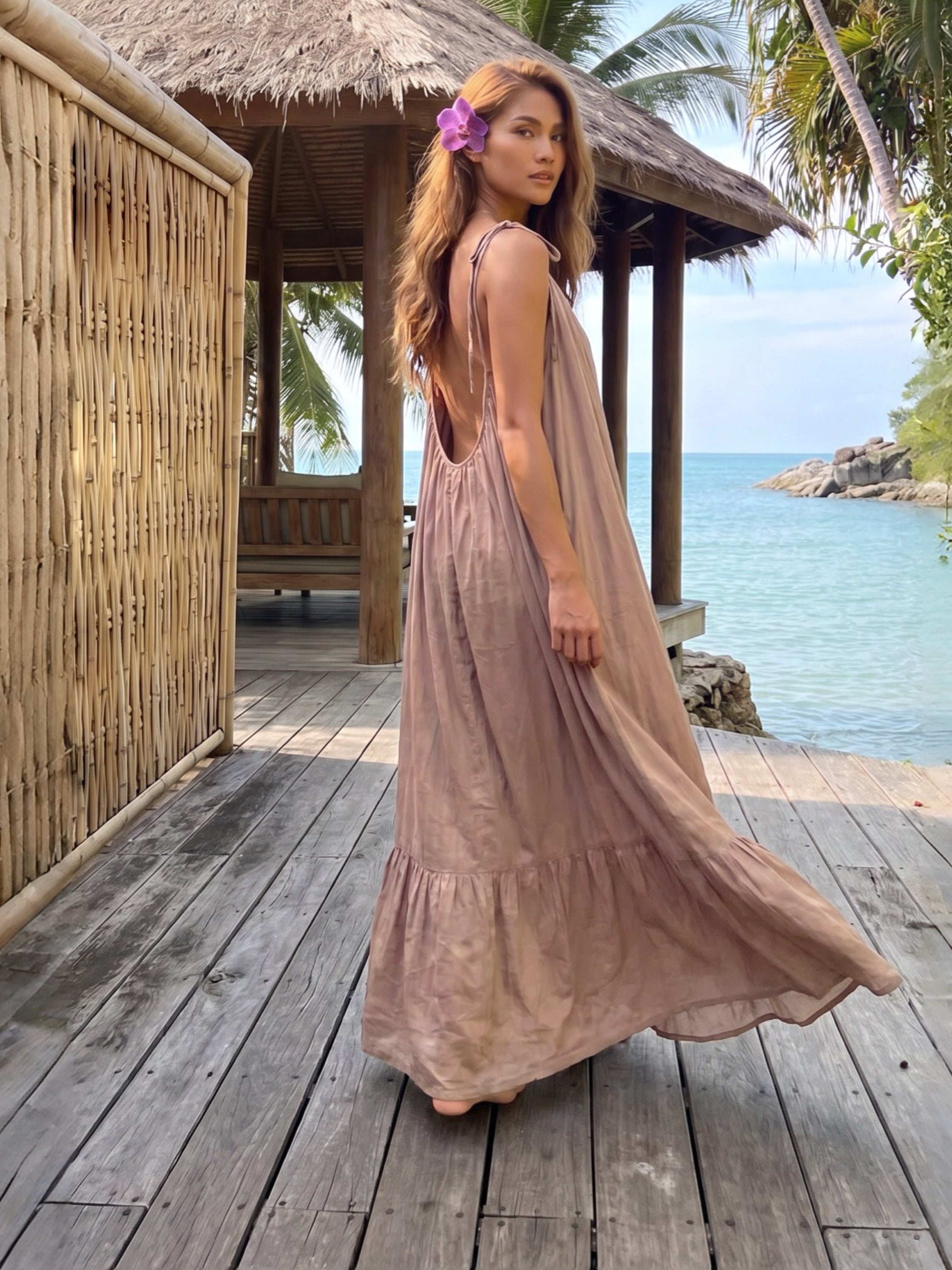 Woman in open back maxi dress in soft nude, handmade backless resort dress with tie shoulders, flowy ruffle hem, and beach vacation style standing on a wooden deck with a thatched-roof structure and ocean view in the background.
