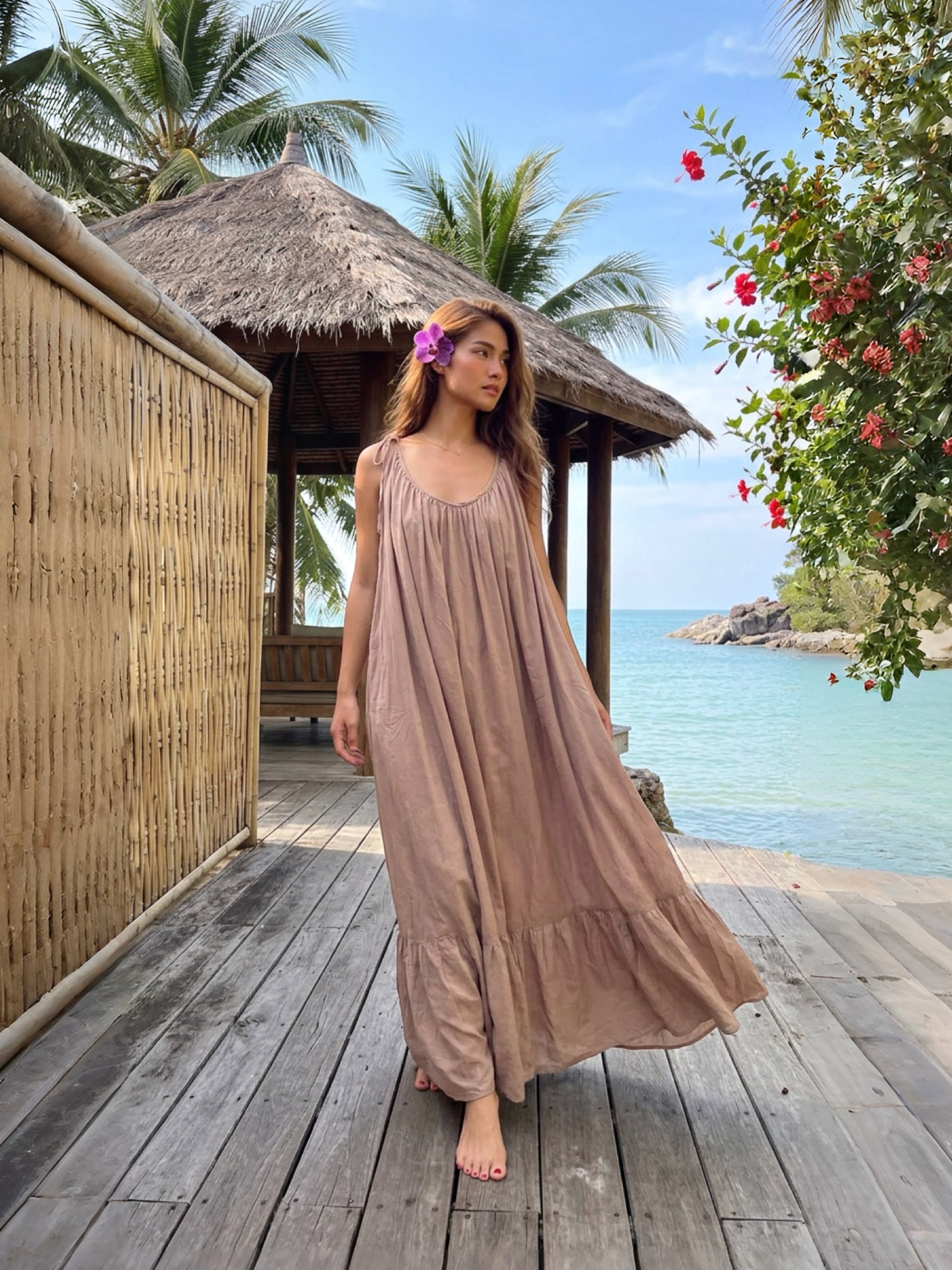 Woman in a long beige open back maxi dress standing on a wooden deck with ocean and tropical setting, perfect resort dress with tie shoulders, flowy ruffle hem, and beach vacation style