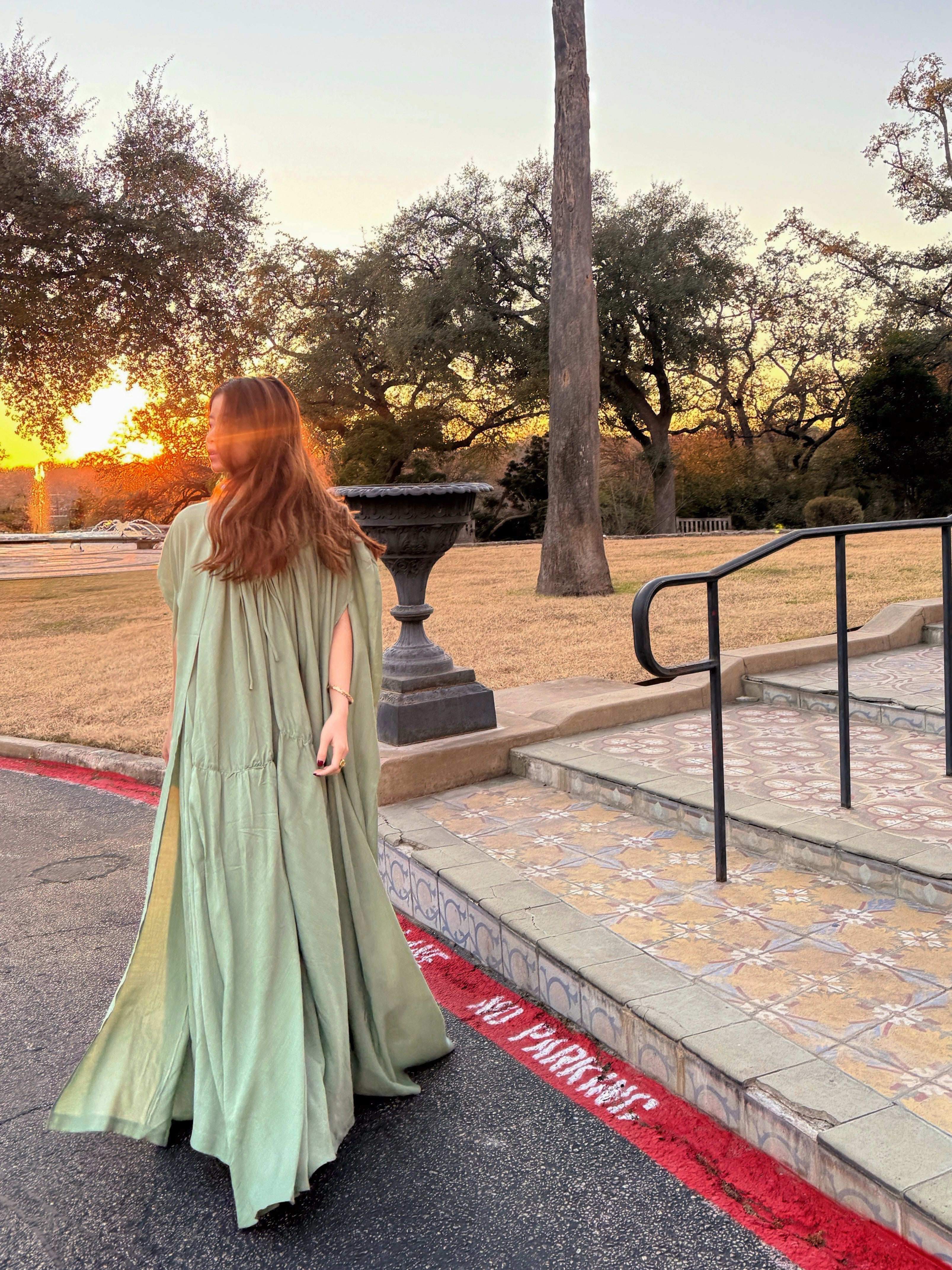 Woman in a sage green maxi dress with cape detail standing in a sunlit hallway, a handmade resort dress and beach wedding guest style by COCO DE CHOM