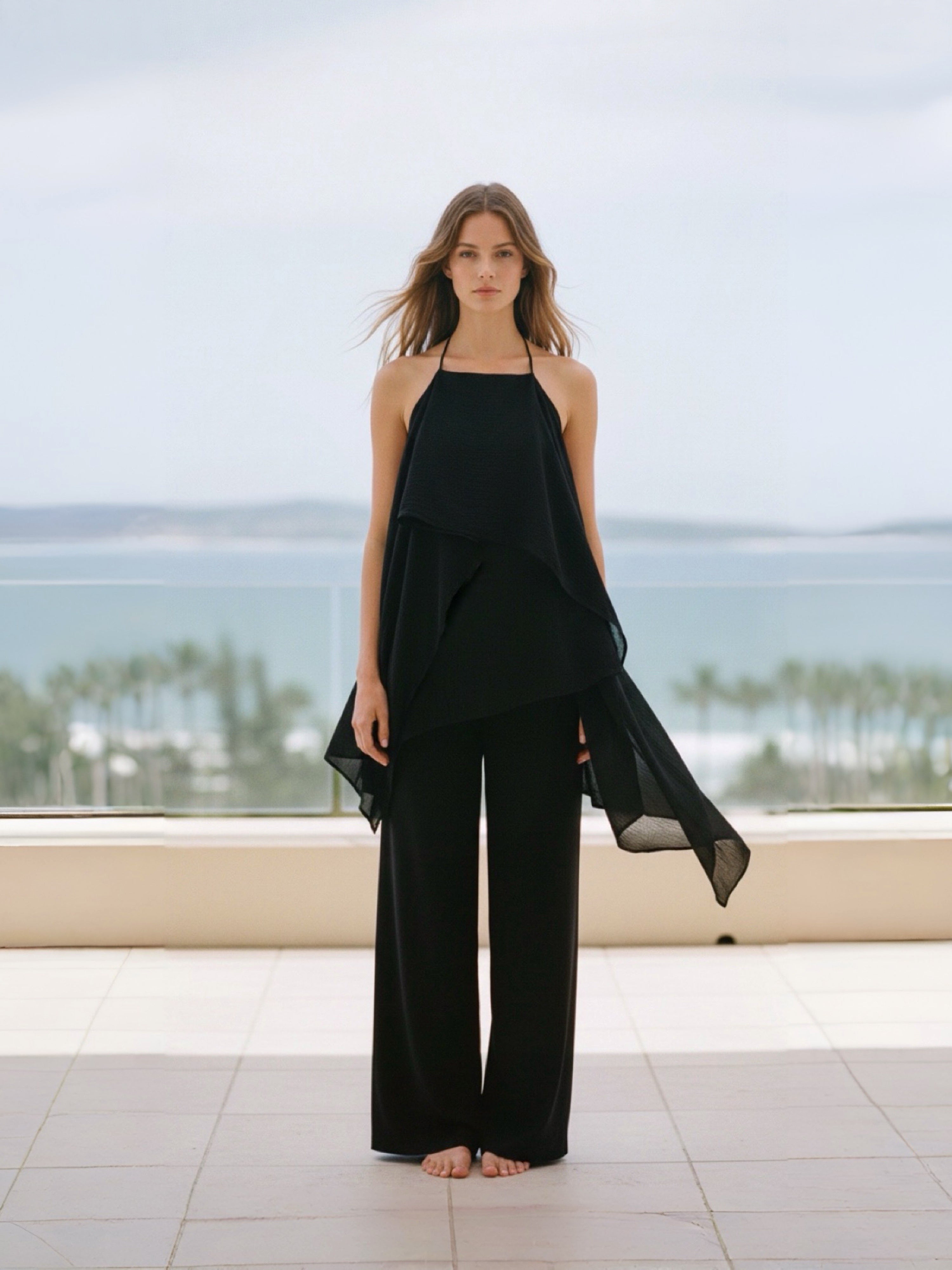 Woman in a black flowy kaftan top standing on a balcony with a scenic view of trees and water.