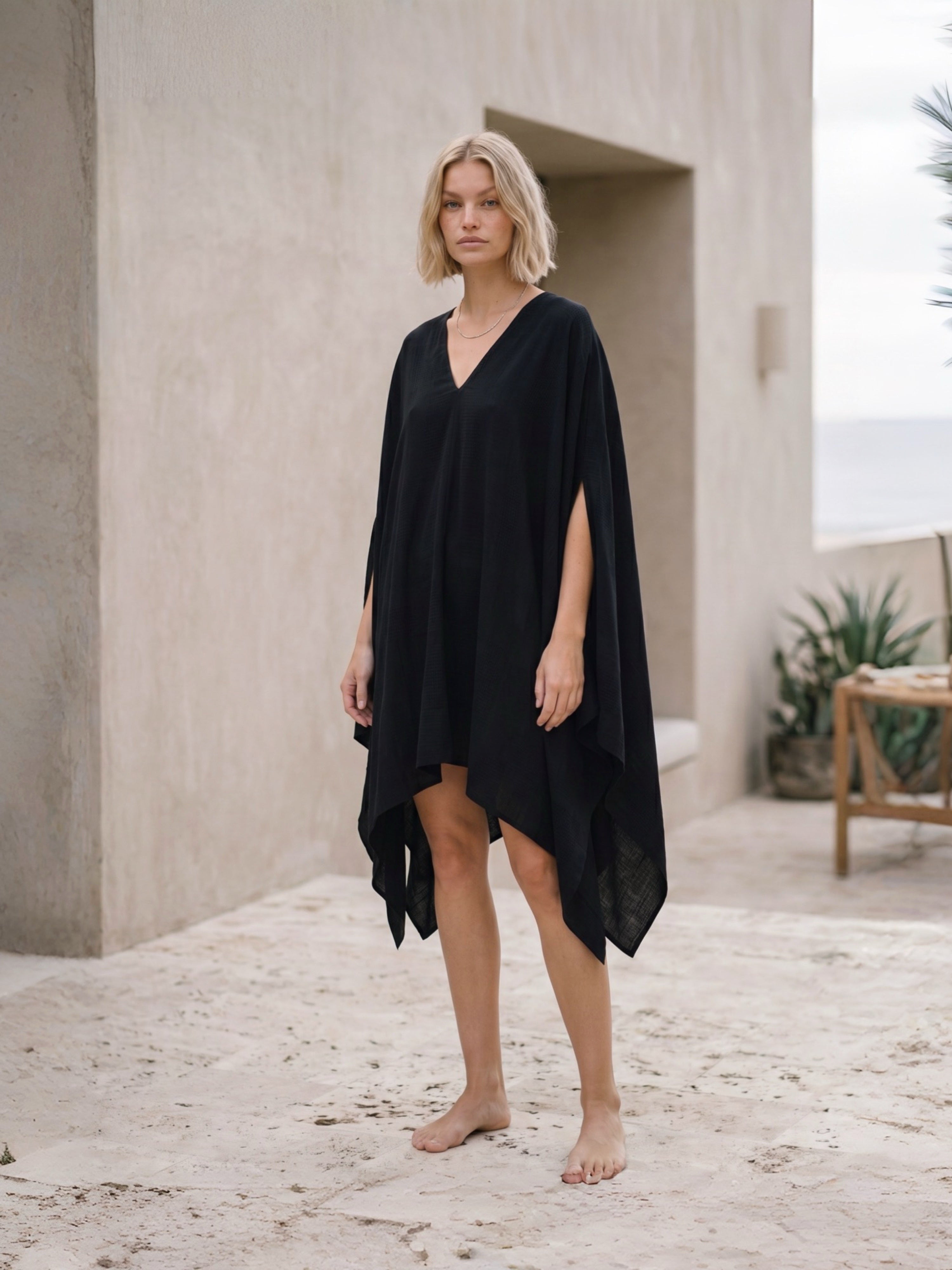 A woman wearing a black Mini Kaftan beach Cover Up, standing barefoot on a sandy patio with a modern concrete wall and outdoor seating in the background.