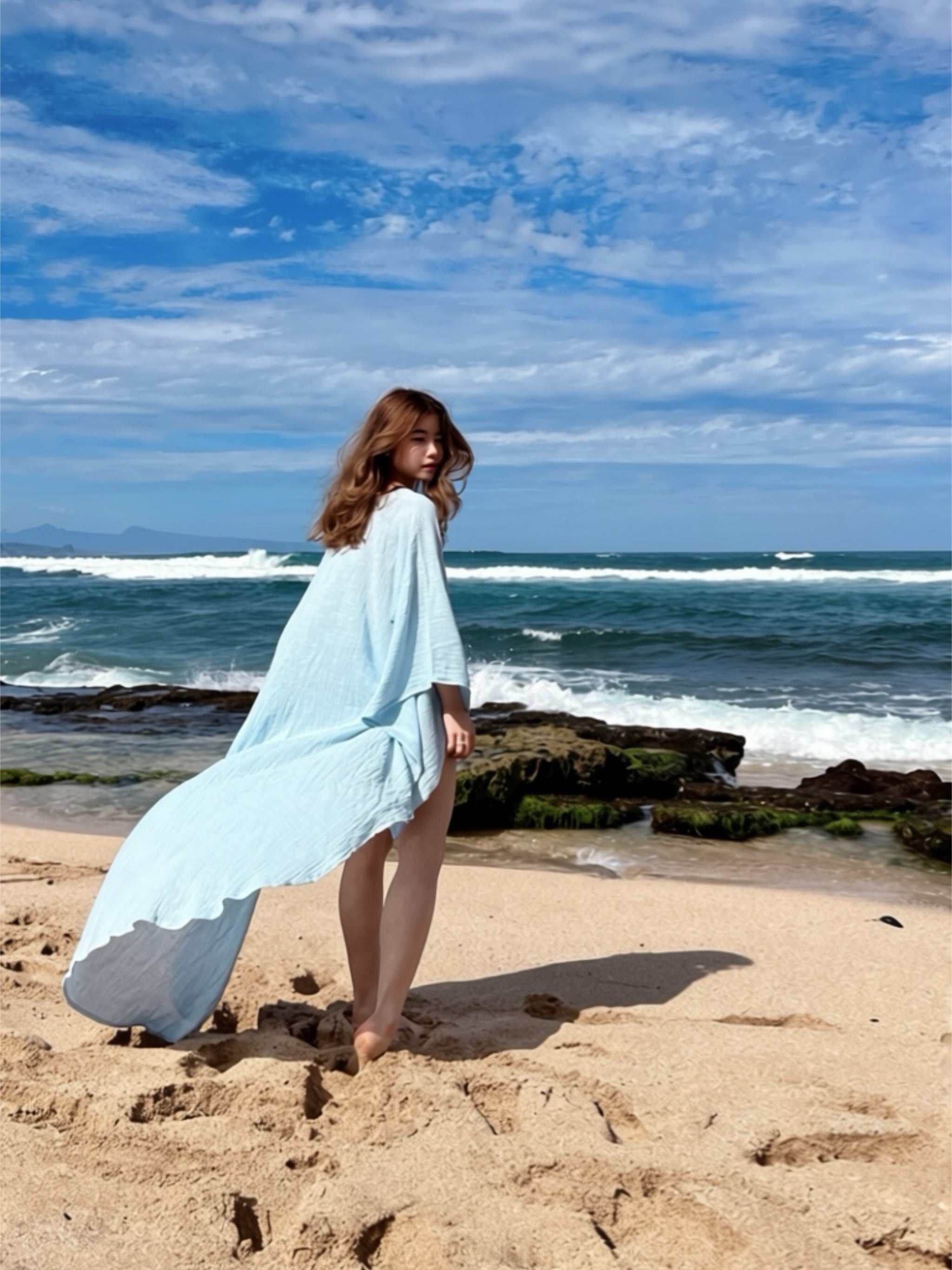 Woman in a light blue linen kaftan top standing on a beach with ocean waves in the background