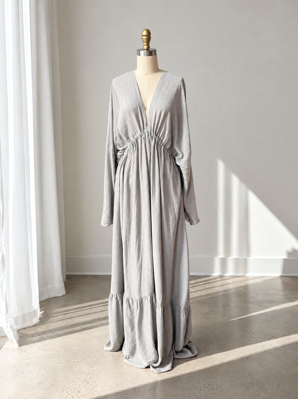 A light gray long-sleeve maxi dress with a V-neckline and gathered waist, displayed on a dress form mannequin in a bright, sunlit room with white curtains and a concrete floor.