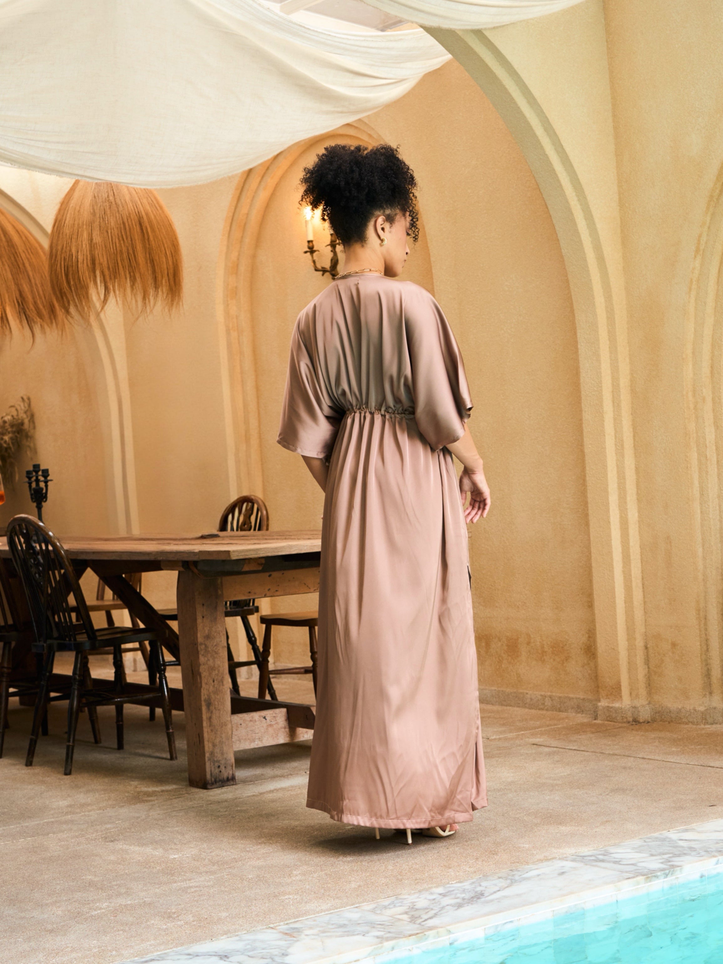Woman wearing a mocha rose kaftan maxi dress with deep V-neck and self-tie waist, styled as resort wear and a beach wedding guest dress by COCO DE CHOM