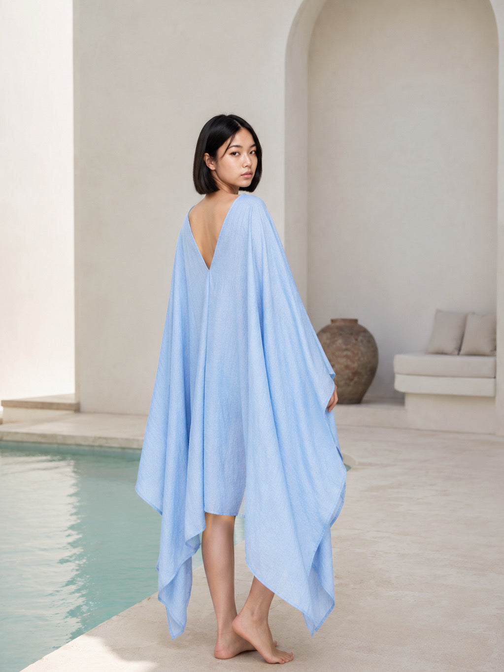 A woman wearing a light blue mini kaftan swim cover-up walks barefoot next to a swimming pool in a minimalist, sunlit worn by the pool, lightweight oversized beach cover up and resort top with deep V neckline for vacation and summer travel