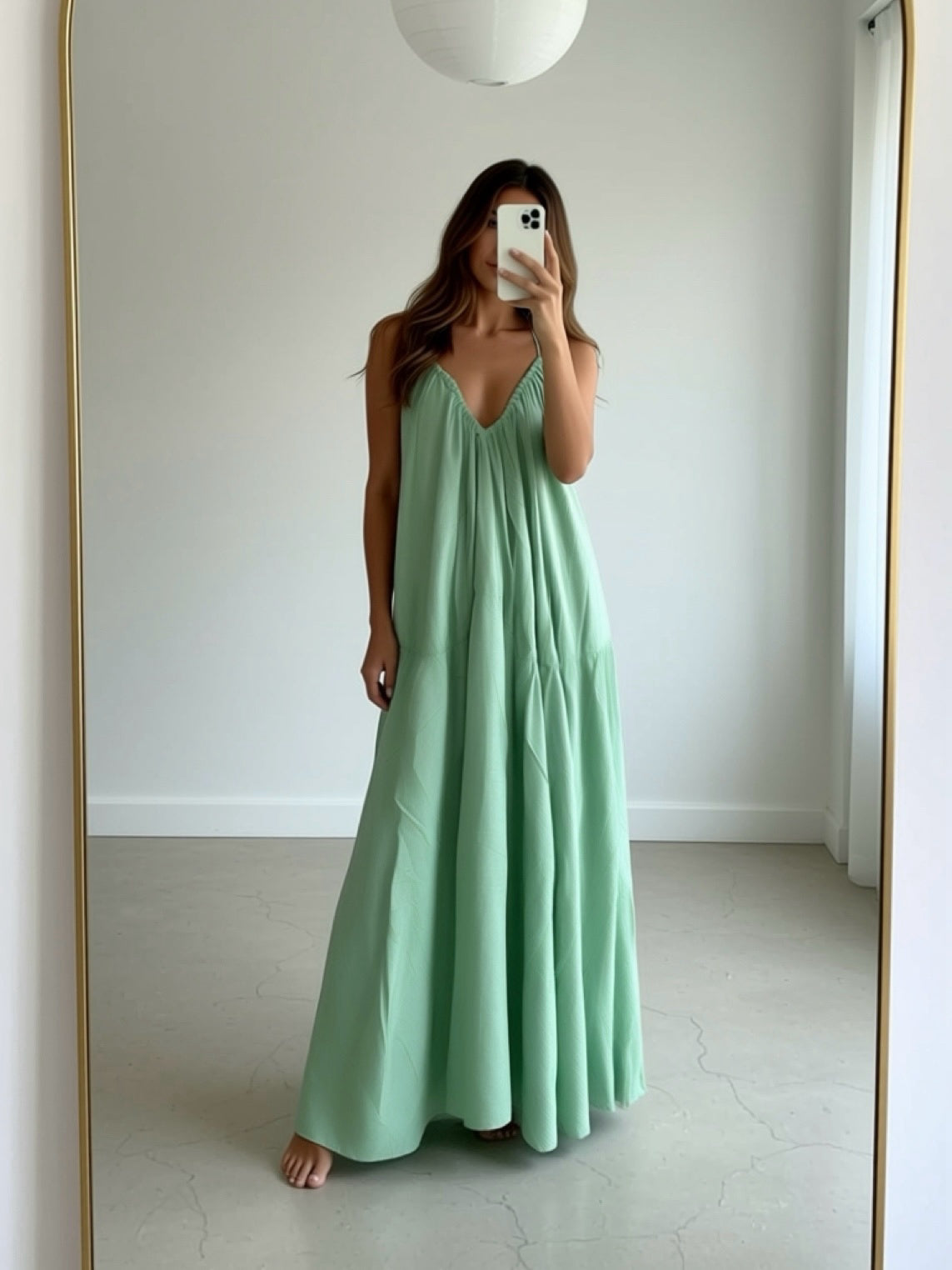 Woman in a sage green maxi dress with cape detail standing in a sunlit hallway, a handmade resort dress and beach wedding guest style by COCO DE CHOM