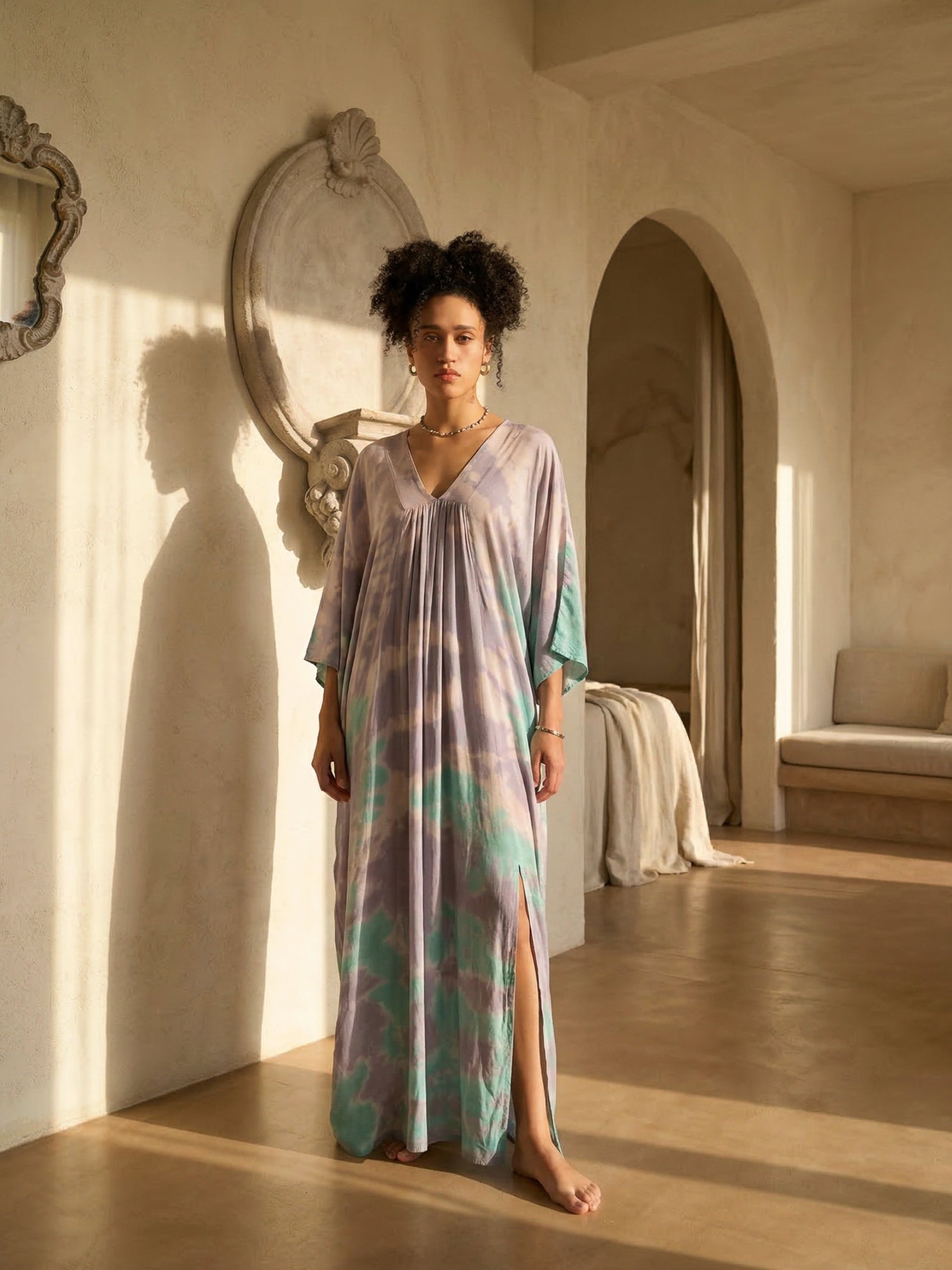 Woman in a long, teal tie dye kaftan dress dress standing in a sunlit room with arched doorways.