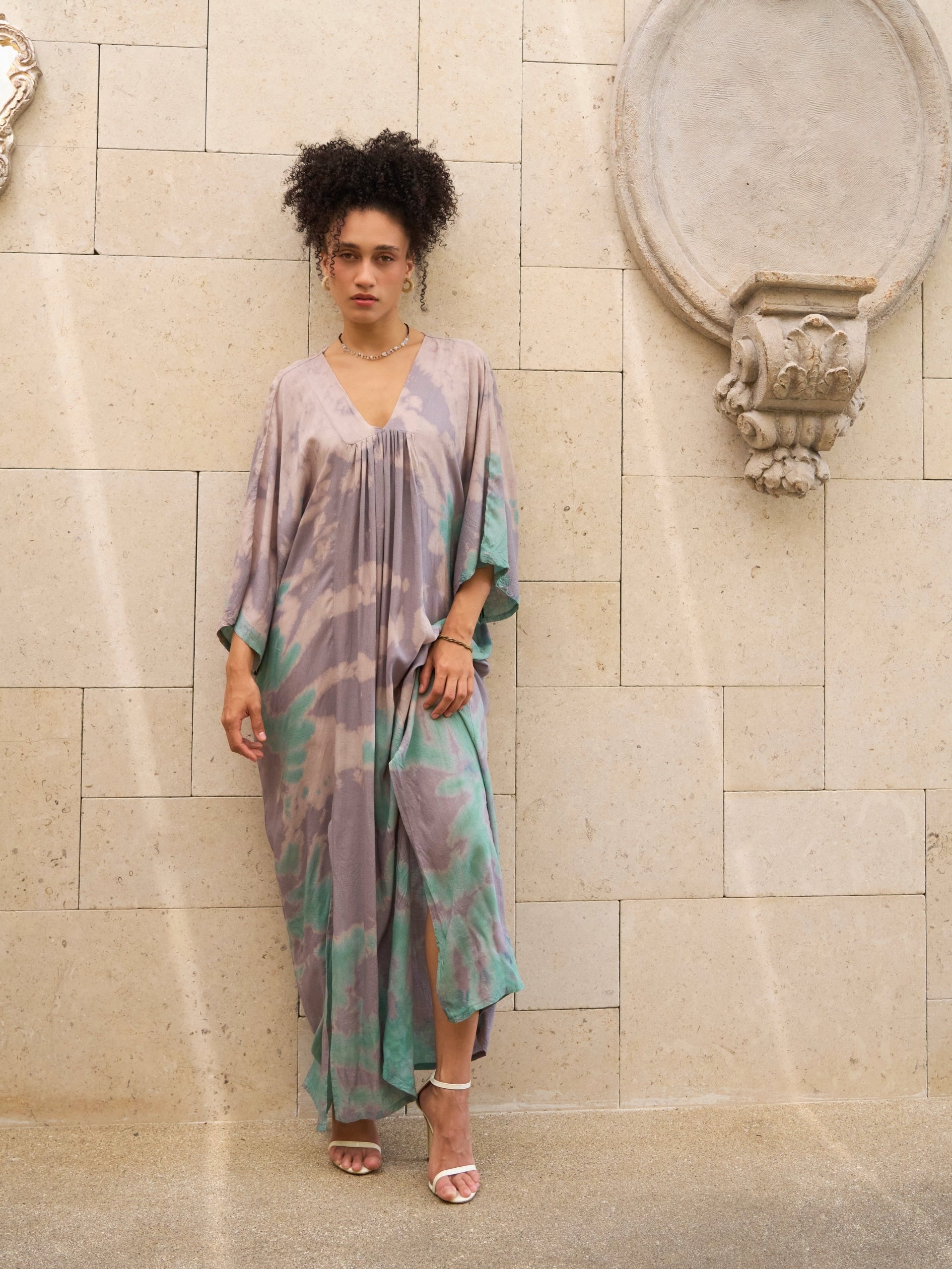 Woman wearing a teal tie-dye kaftan dress , goddess dress 
standing against a stone wall.