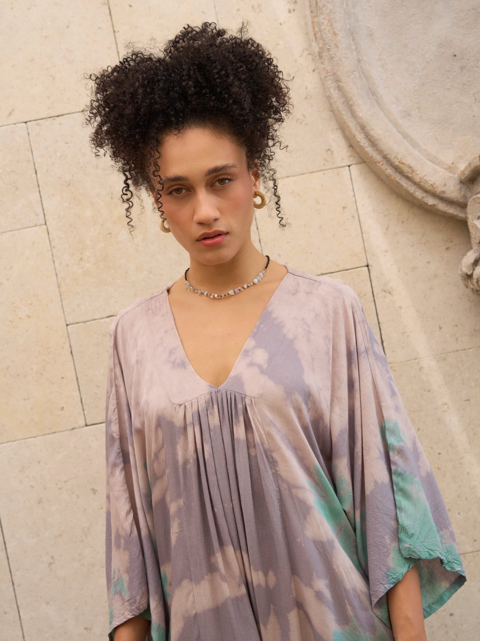 Woman wearing a Goddess tie-dye kaftan dress against a stone wall.