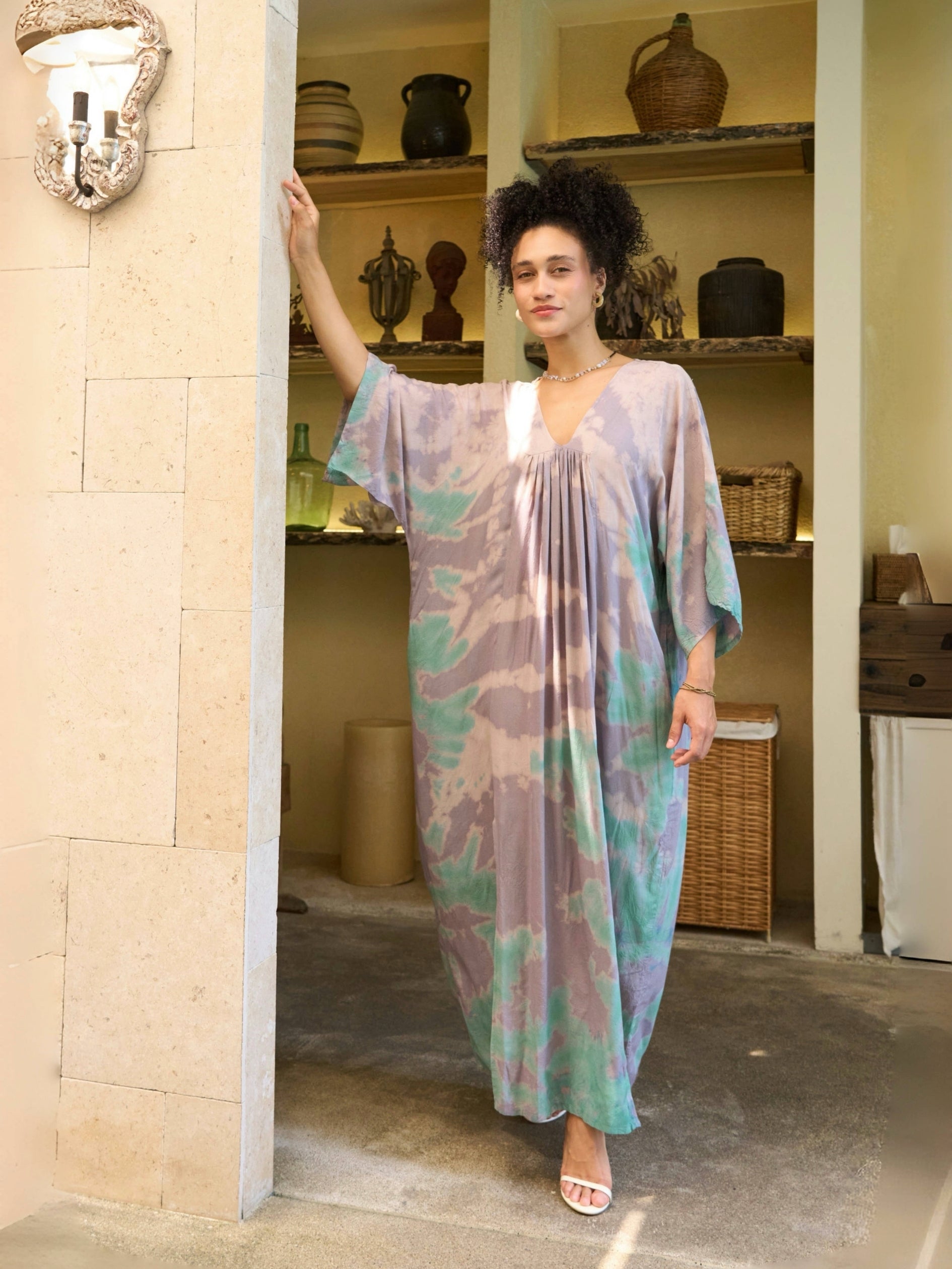 Woman wearing a teal tie-dye kaftan dress standing in a room with shelves and decor.