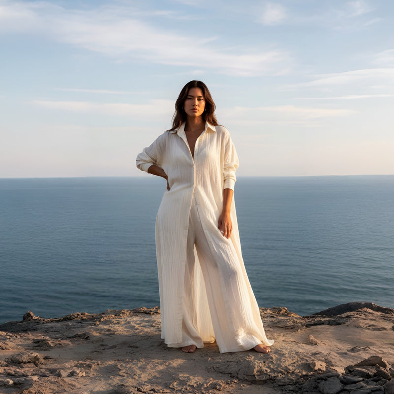 Woman in a natural cotton maxi shirt dress  with white cotton wide leg pants standing on a cliff overlooking the ocean.