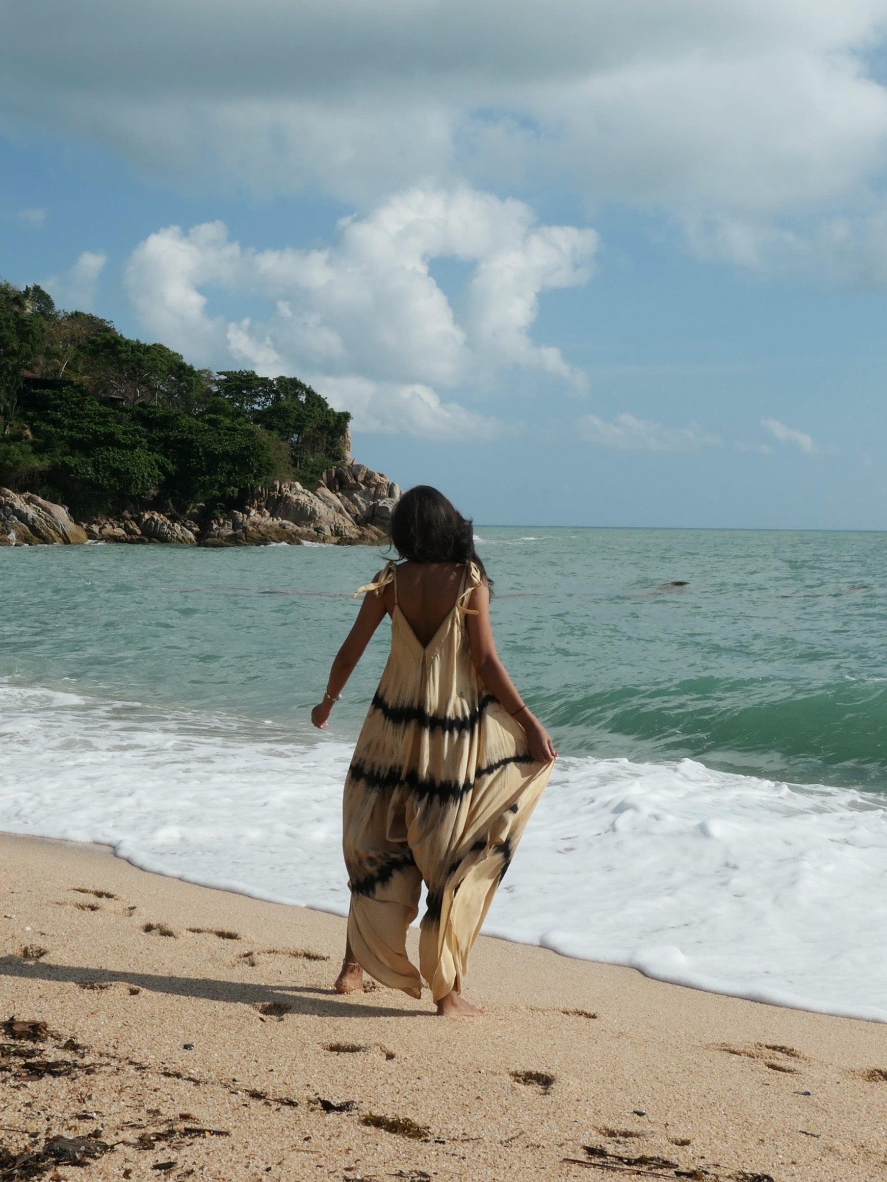 Woman in tie dye jumpsuit  with an oversized fit and back-V silhouette, perfect for yoga, beach cover-up, or casual day-to-night wear by Coco De Chom standing on a sandy beach with ocean and rocks in the background. 