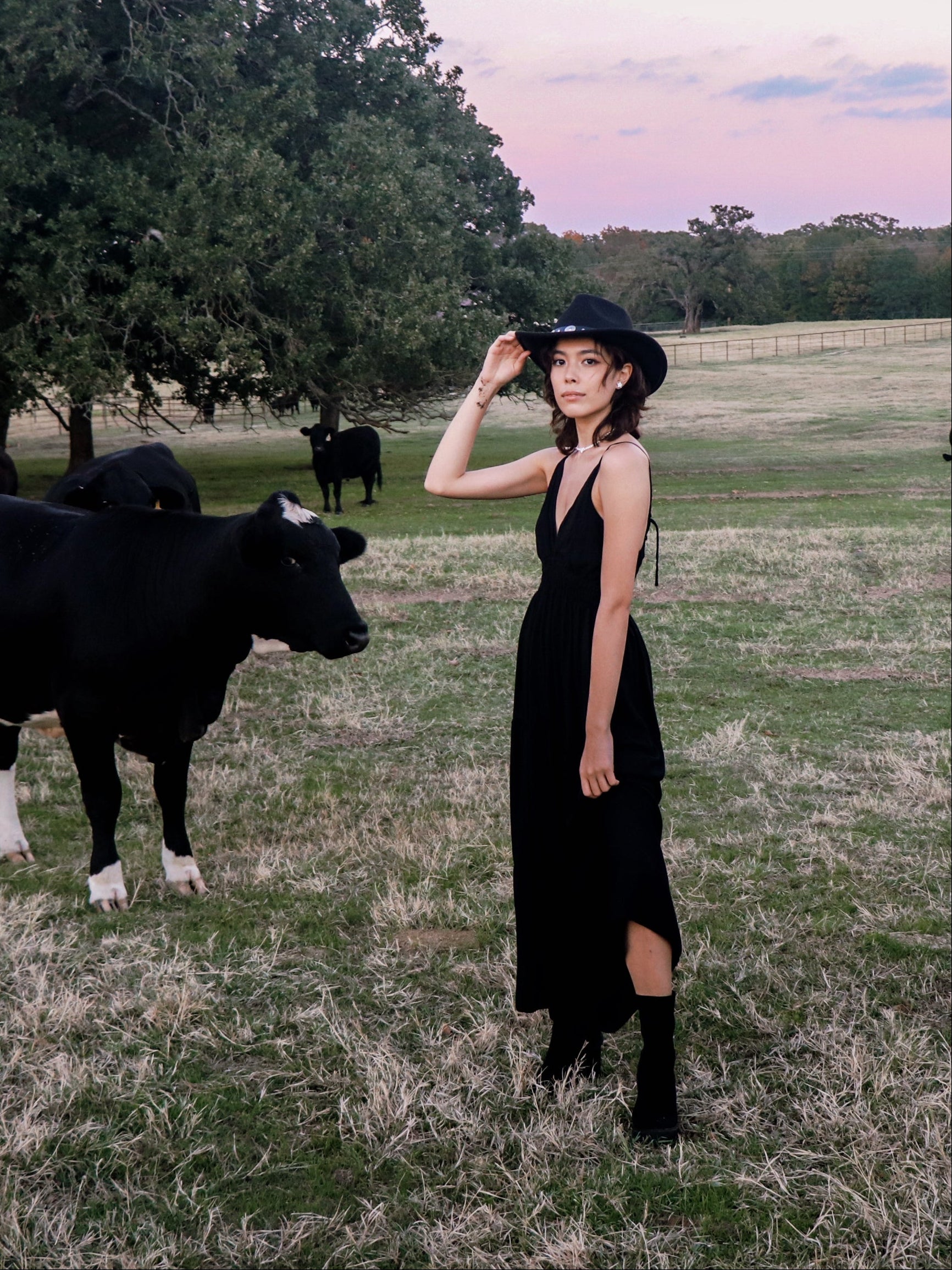A woman standing in a field with a cow in the background, wearing a black maxi dress with a smocked bodice.