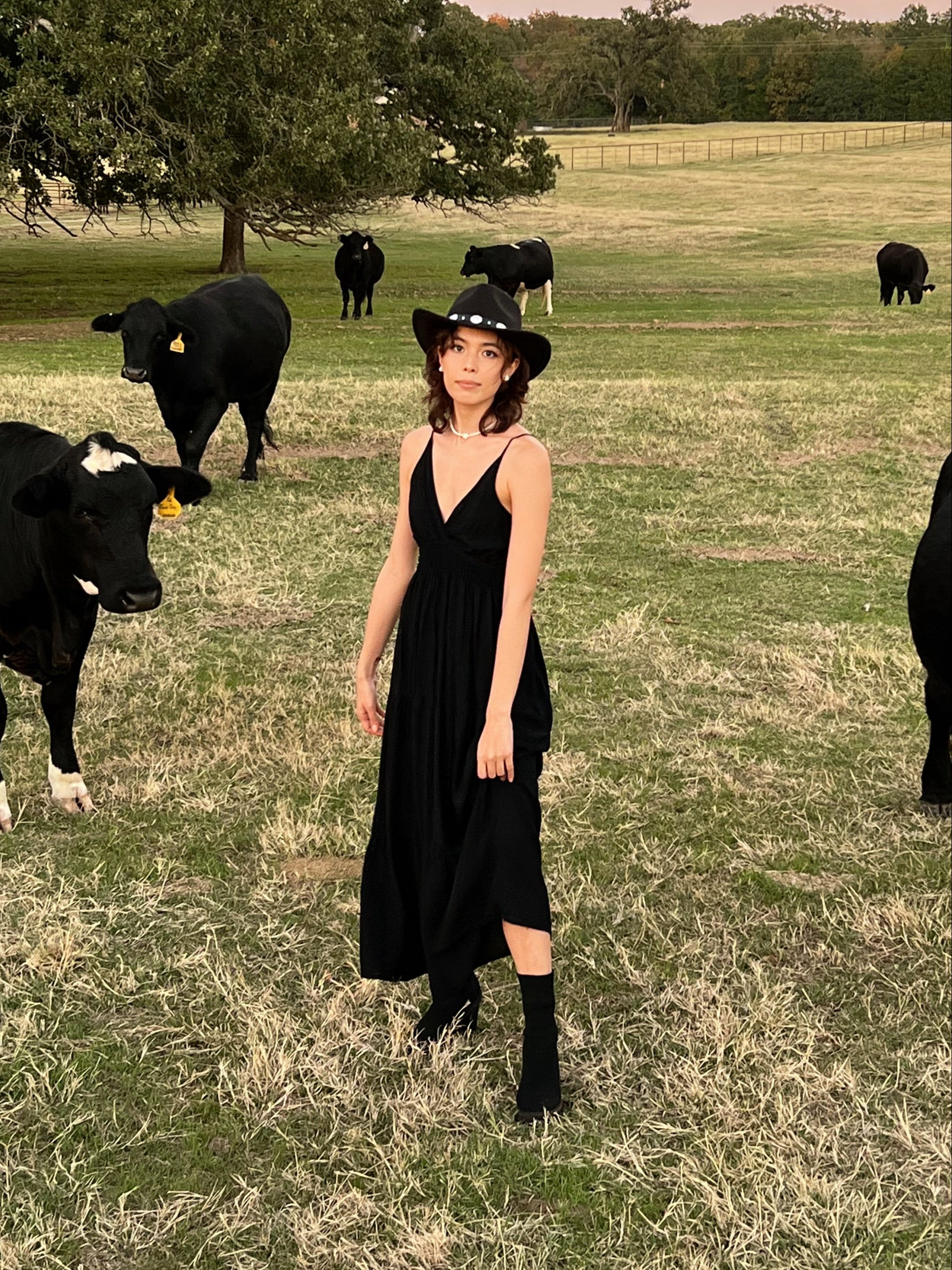 A woman standing in a field with a cow in the background, wearing a black maxi dress with a smocked bodice.