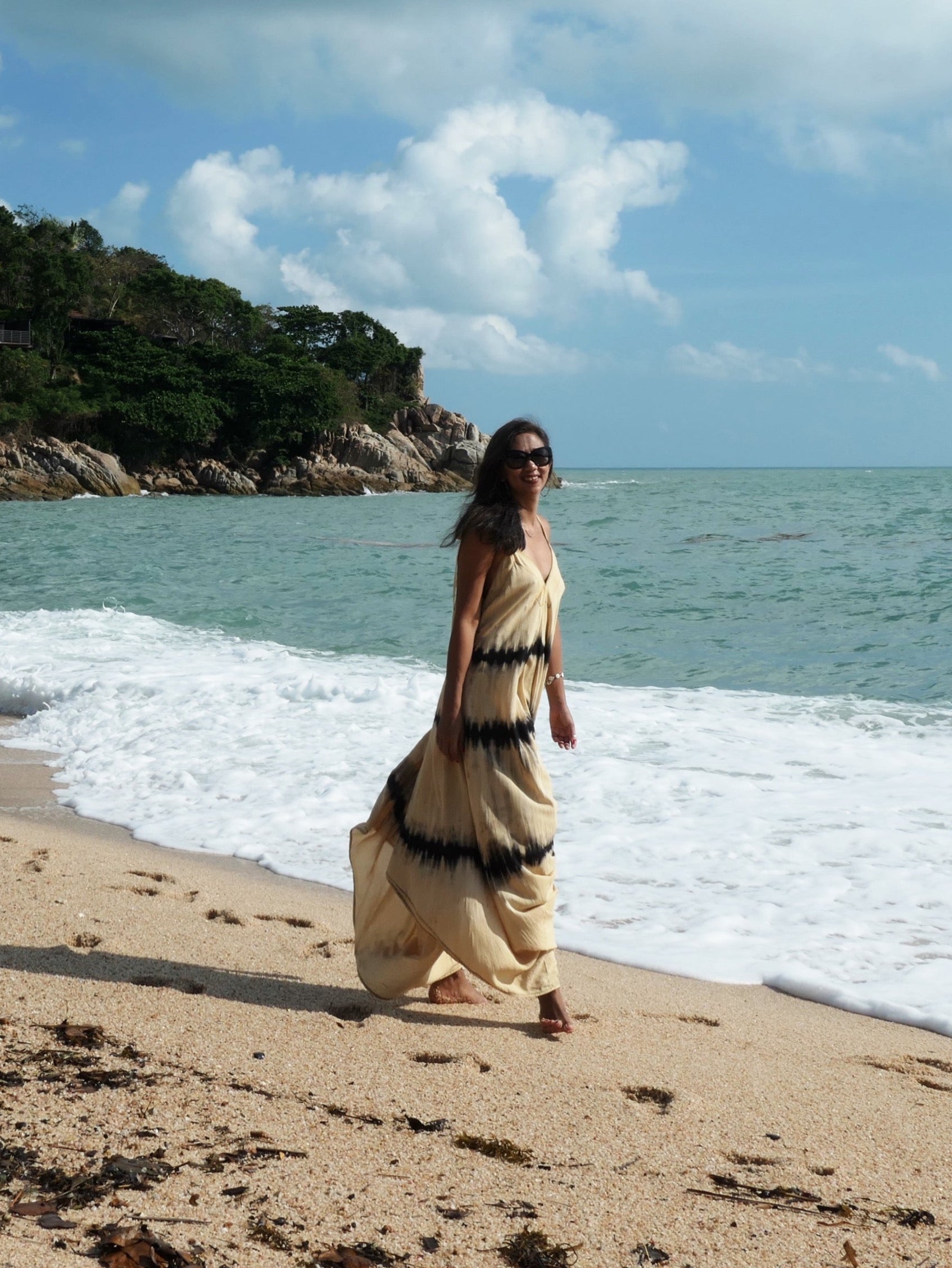 Woman in tie dye jumpsuit  with an oversized fit and back-V silhouette, perfect for yoga, beach cover-up, or casual day-to-night wear by Coco De Chom standing on a sandy beach with ocean and rocks in the background. 